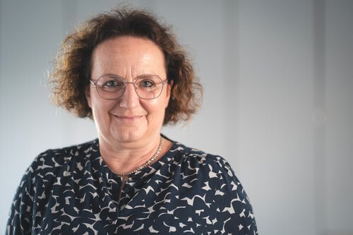 Barbara Kl&auml;sle mit lockigem, schulterlangem Haar tr&auml;gt eine schwarze Bluse mit wei&szlig;em Muster.