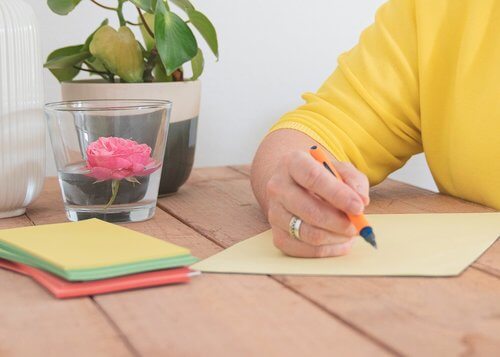 Hand mit orangem Stift schreibt auf gelbes Papier, daneben Stapel bunter Notizzettel und Glas mit rosa Rose im Wasser auf Holztisch.